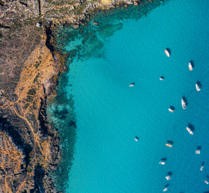 Draufsicht auf die K&uuml;ste Siziliens mit t&uuml;rkisblauem Wasser und zahlreichen Booten entlang der felsigen Uferlinie.