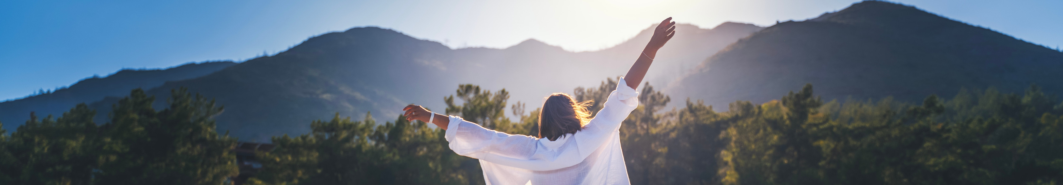 Eine Frau in einem wei&szlig;en Hemd genie&szlig;t mit erhobenen H&auml;nden die Morgensonne vor der Kulisse der Berge bei Sonnenaufgang