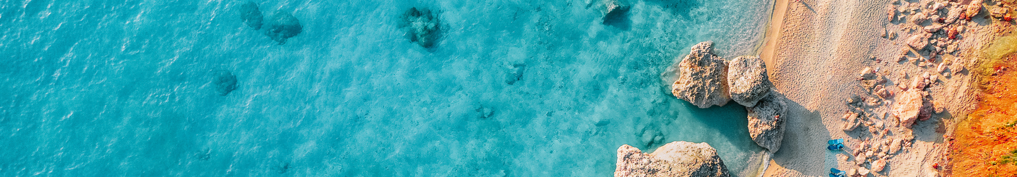 Drohnenaufnahme eines Strandes mit t&uuml;rkisblauem Wasser, Felsen im Sand und Reihen von Liegest&uuml;hlen.