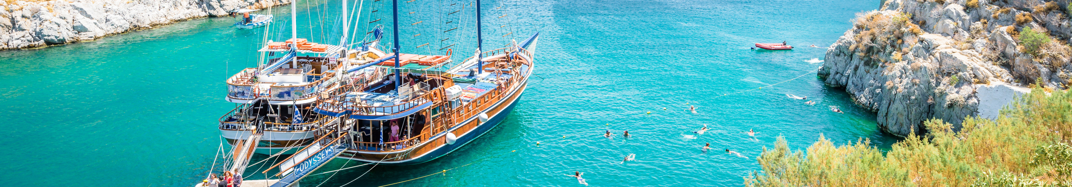 Badebucht mit türkisblauem Wasser, Felsen, Badenden und zwei traditionellen Segelbooten am Steg.