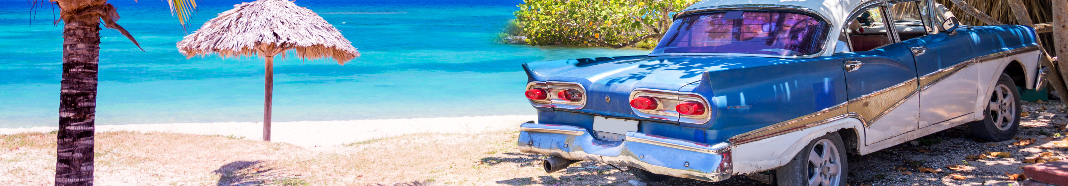 Ein klassisches Oldtimer-Auto steht im Schatten von B&auml;umen an einem tropischen Strand in Kuba, neben einer Palme und einer Strohh&uuml;tte, mit t&uuml;rkisblauem Meer im Hintergrund.