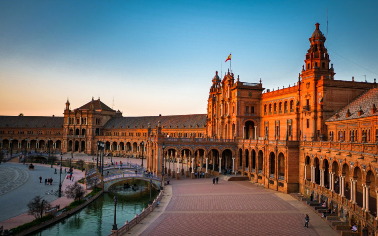 Panoramablick auf die Plaza de España in Sevilla bei warmem Abendlicht, mit halbrundem Backsteingebäude, Arkadengängen, Brücken über einen Kanal und blauem Himmel.