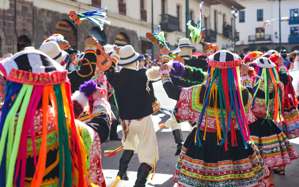 Teilnehmende des Inti Raymi-Festes in traditioneller Kleidung tanzen und musizieren auf einer engen Straße, geschmückt mit bunten Bändern und Instrumenten, während Zuschauer das Spektakel verfolgen.