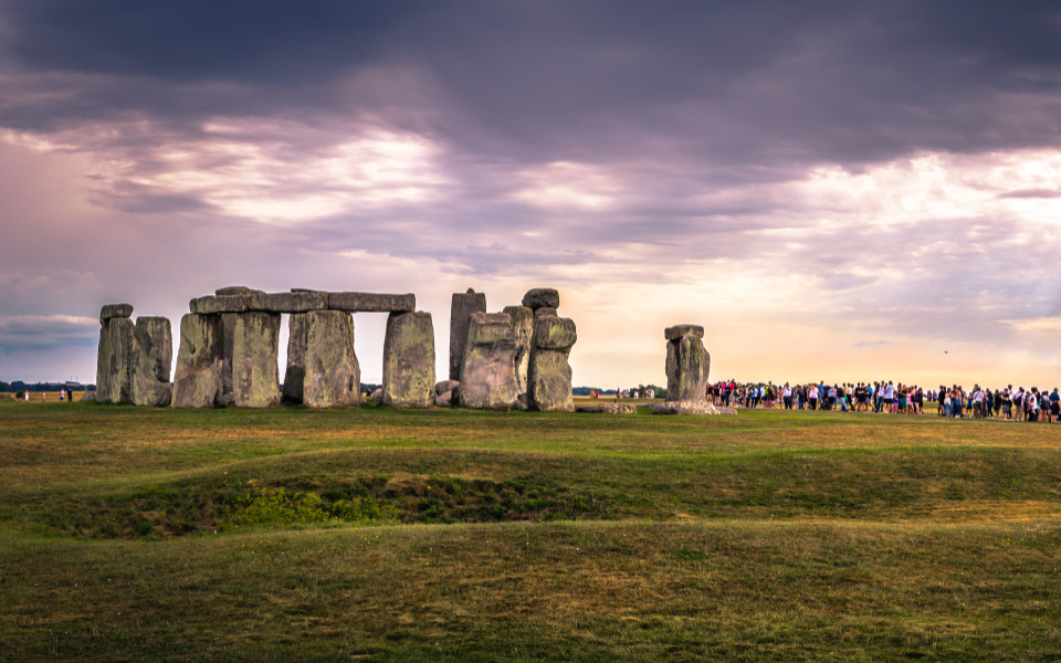 Panoramablick auf Stonehenge unter einem dramatischen, bewölkten Himmel, eine Gruppe von Besuchern betrachtet das historische Monument aus einiger Entfernung.
