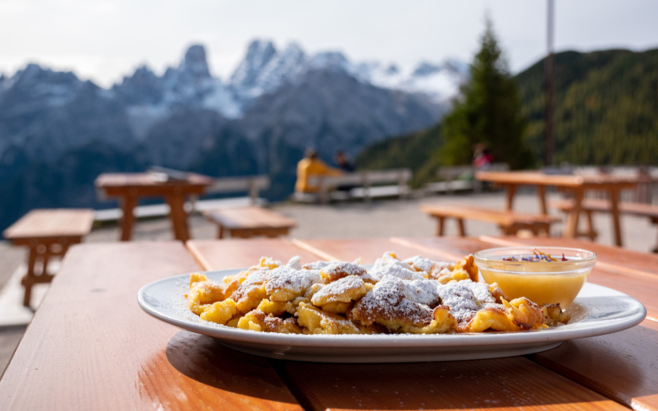 Ein Teller Kaiserschmarrn mit Puderzucker und Apfelmus wird auf einem Tisch serviert, im Hintergrund sind Berge und B&auml;nke zu sehen.