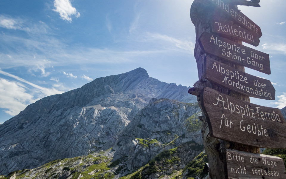 Wegweiser mit mehreren Holzschildern vor der markanten Felswand der Alpspitze in Bayern bei strahlend blauem Himmel