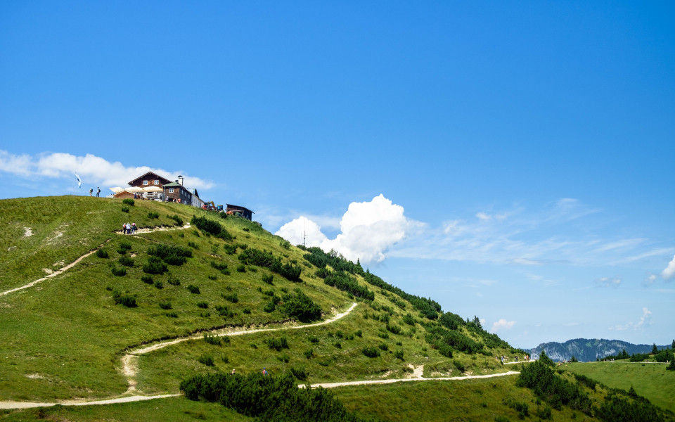 Wanderweg f&uuml;hrt den gr&uuml;nen Hang des Wankbergs hinauf zu einer Bergh&uuml;tte, unter strahlend blauem Himmel mit wenigen Wolken.